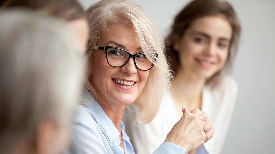 Smiling aged businesswoman in glasses looking at colleague at team meeting, happy attentive female team leader listening to new project idea, coach mentor teacher excited by interesting